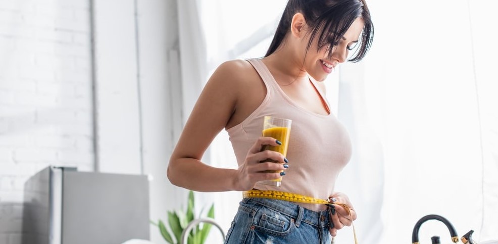 szczupła kobieta na redukcji mierzy obwód talii i trzyma dietetyczne smoothie